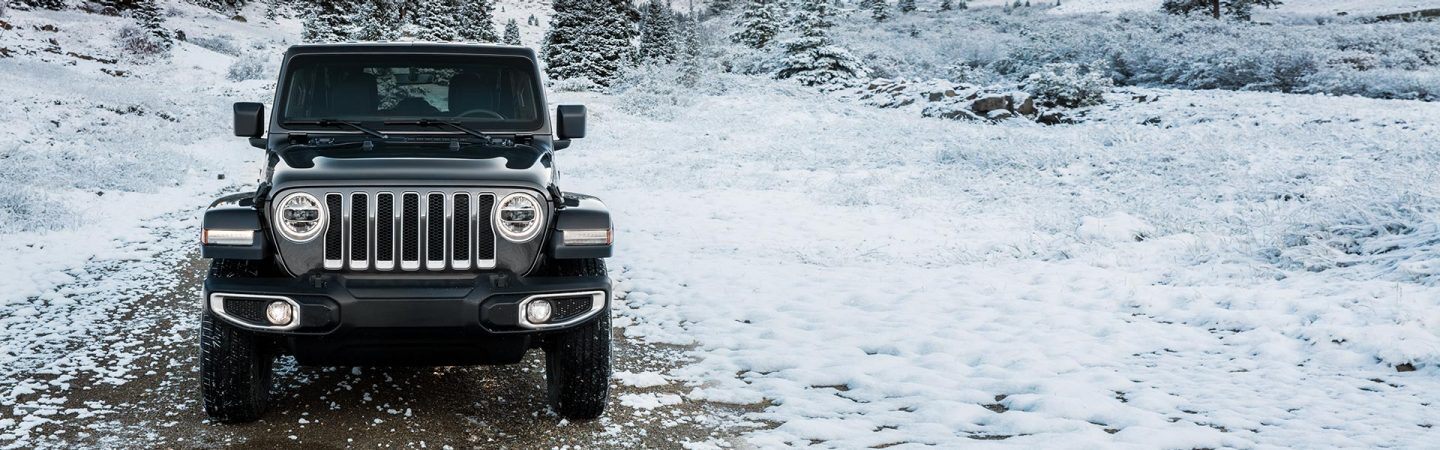 A head-on front view of a 2021 Jeep Wrangler Sahara Unlimited on a snow-covered trail in a wooded area of hills.